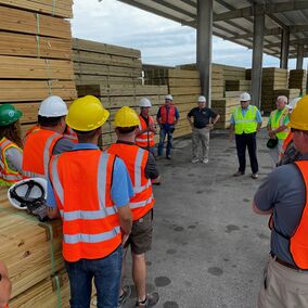 Group of employees gathered under a lumber rack in a lumber yard, wearing safety vests and hard hats